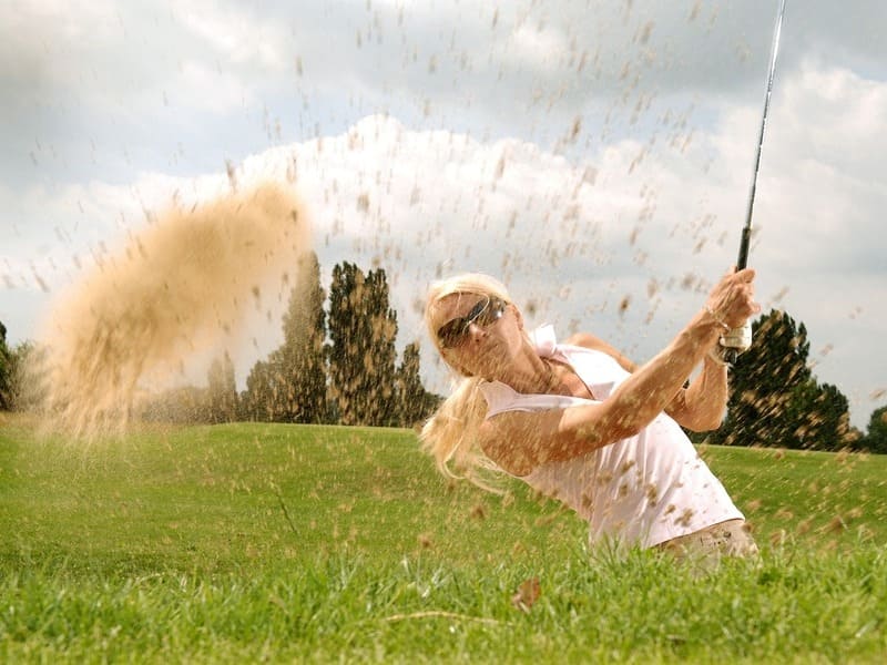mujer golfista rubia y con gafas de sol concluyendo un swing desde un bunker. Arena en el aire. De la mujer asoma sólo medio cuerpo fuera del bunker. Al fondo cielo nuboso