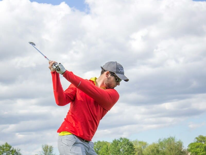 hombre golfista iniciando un swing. Lleva sweater rojo, gorra azul, pantalones grises. Al fondo, arboleda, con predominio de cielo, que está nublado