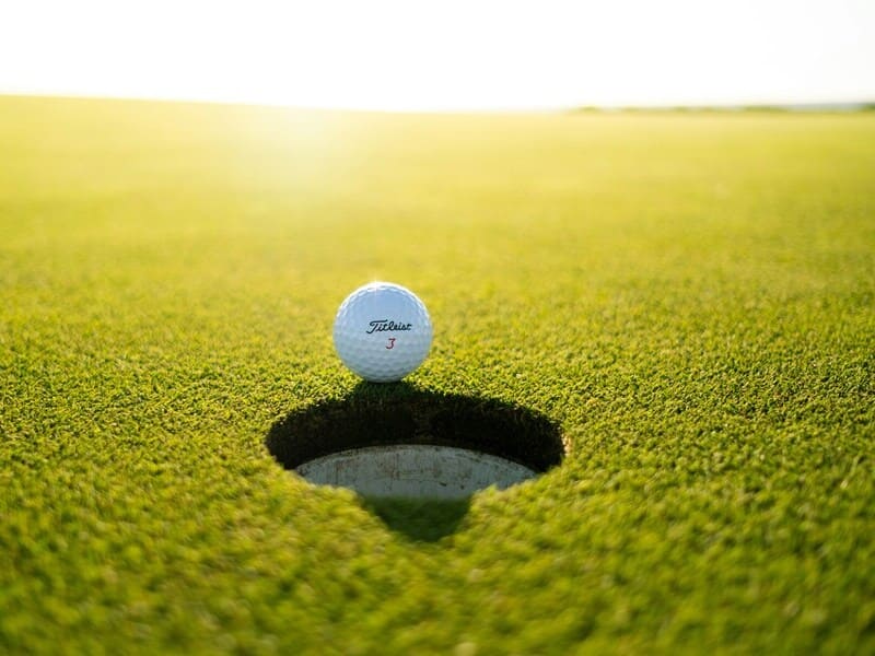 pelota de golf en el filo de un hoy, pero sin caer. La mayoría de la imagen es césped, con un horizonte bañado por la luz del sol