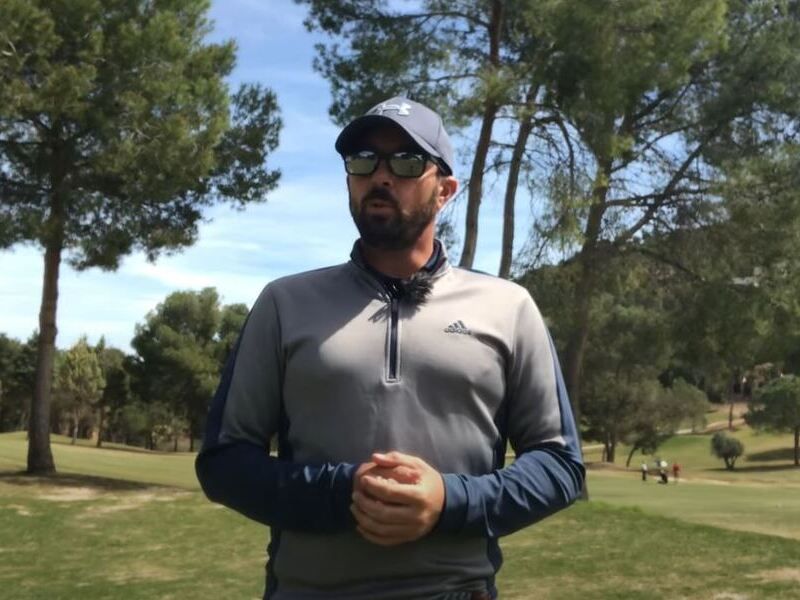 imagen de golfista ataviado con prendas de color grisáceo-azulado, gorra y gafas de sol. Tiene barba. Se ve sólo el torso. Tiene las manos entrelazadas a la altura del abdomen. Al fondo arboleda y cielo casi despejado