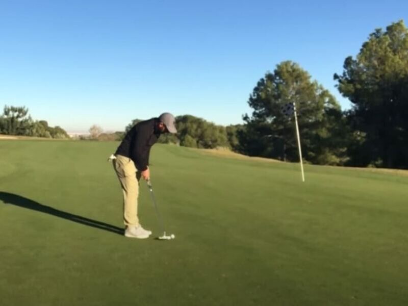 imagen de Rafa Pérez ejecutando golpe de aproximación al hoyo en el green. Viste pantalones beige, sweater azul marino y gorra gris. A la derecha bandera del hoyo. Césped, arboleda y cielo despejado completan la escena.