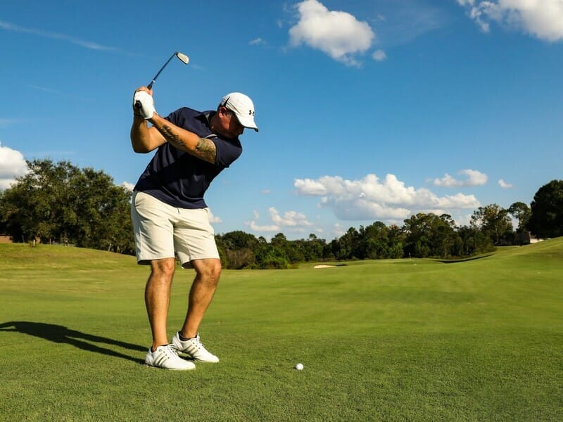 golfista iniciando swing, con pantalones cortos blancos y camisa oscura. Cielo despejado y arboleda al fondo