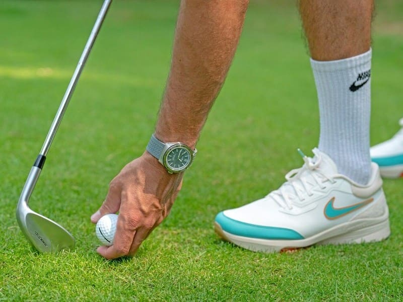 mano de golfista con reloj recogiendo bola del césped. Se ve también algo de su zapatilla y su palo