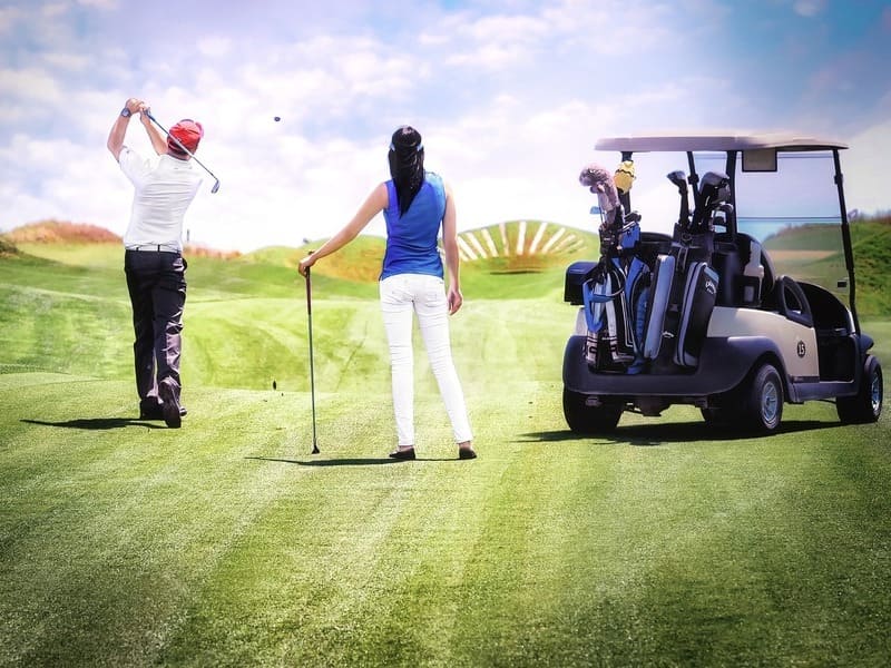 imagen de mujer y hombre golfistas de espaldas sobre el césped. Un carrito eléctrico a su derecha. Todos sobre césped. Al fondo, cielo con nubes y claros
