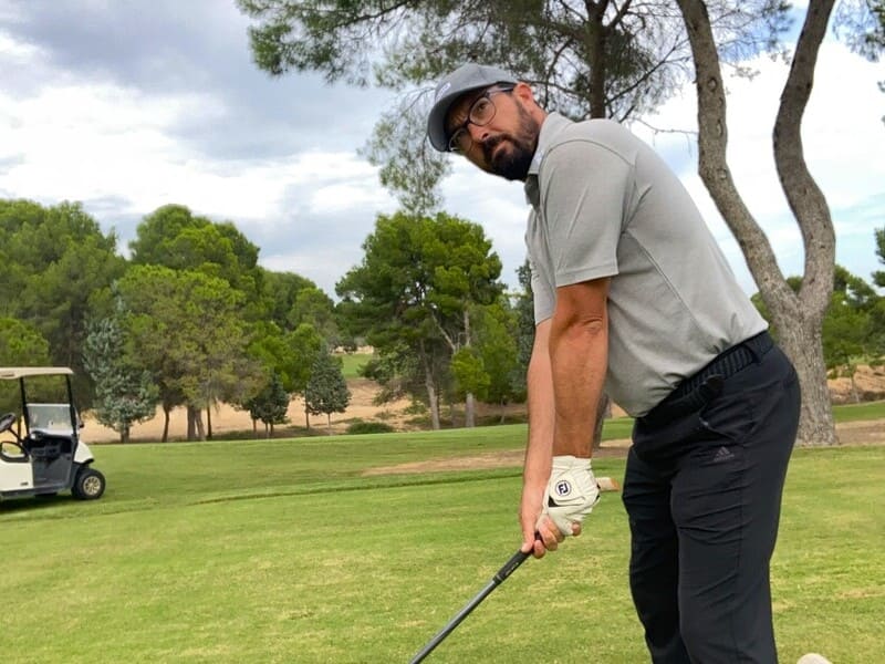 imagen de rafa pérez (golfista), en posición previa a swing, mirando el hoy. Lleva gorra azul, camisa gris y pantalones azul marino. Al fondo césped, arboleda y coche de golf blanco.