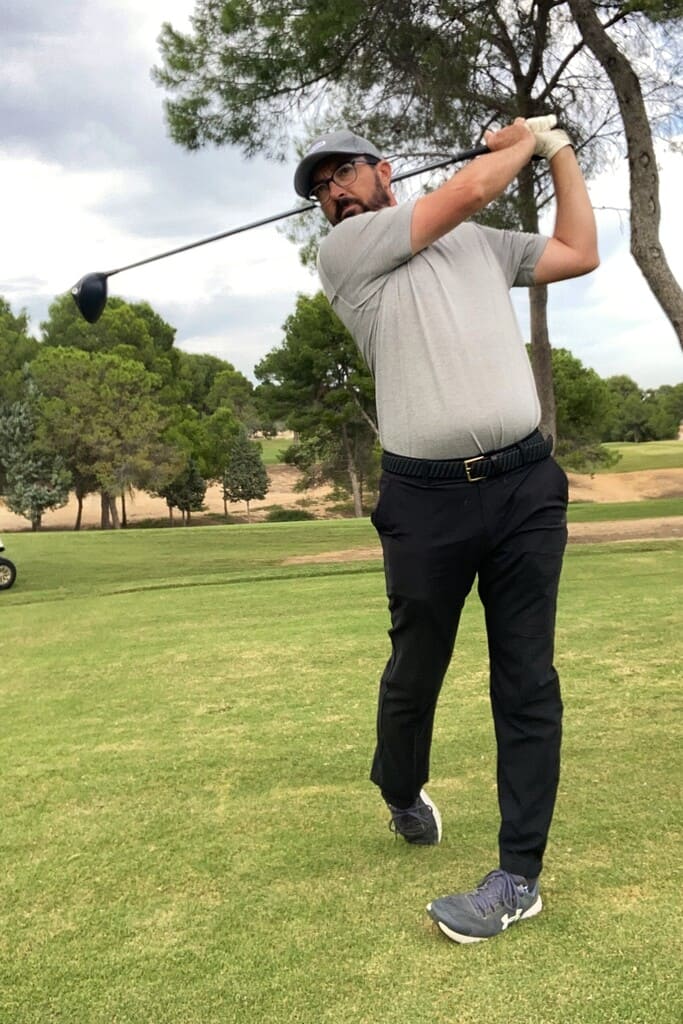 Foto de Rafa Pérez (golfista) ejecutando un swing en posición final. Lleva gorra azul grisáceo, gafas, camiseta gris y pantalones azul marino. Está sobre césped, con algunos árboles al fondo. El cielo, con nubes y claros.