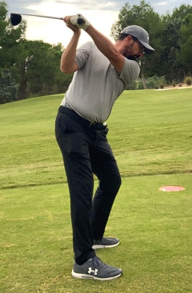foto de rafa pérez, golfista, iniciando la ejecución de un swing. césped alrededor, arboleda al fondo y cielo con nubes y claros. Viste pantalones azul marino y camisa gris. Lleva gorra